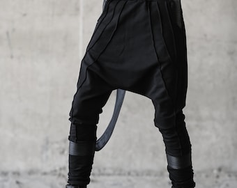 May include: Black harem pants with pleated details and a drop crotch. The pants have a wide waistband and are paired with black boots. A black strap is attached to the pants. The outfit is all black.