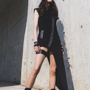 May include: A black sleeveless dress with a high collar and a thigh-high slit. The dress features leather accents and is paired with black boots and sunglasses. A garter belt and wrist cuffs complete the edgy look.