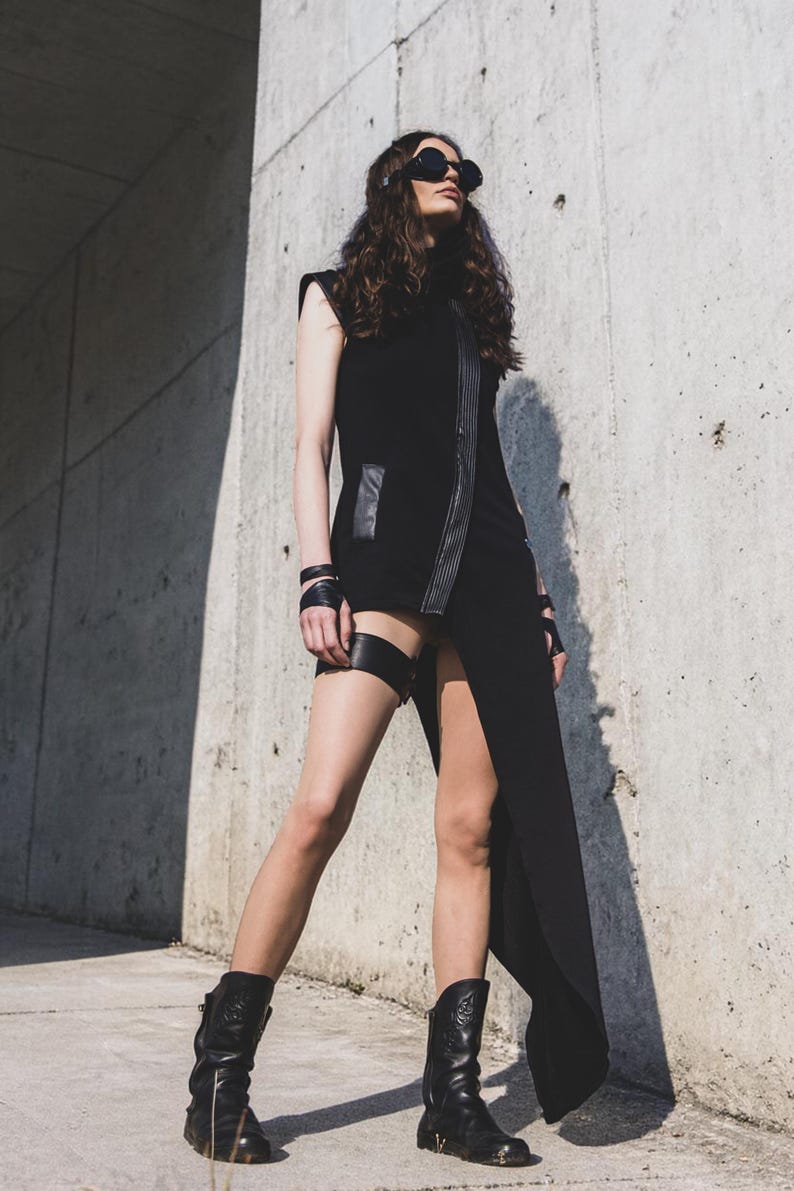May include: A black sleeveless dress with a high collar and a thigh-high slit. The dress features leather accents and is paired with black boots and sunglasses. A garter belt and wrist cuffs complete the edgy look.