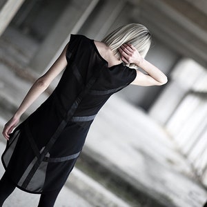 May include: Black dress with sheer panels and geometric accents. The dress features a sleeveless design with a high neckline and a flowing, asymmetrical hemline. The dress is paired with black leggings.