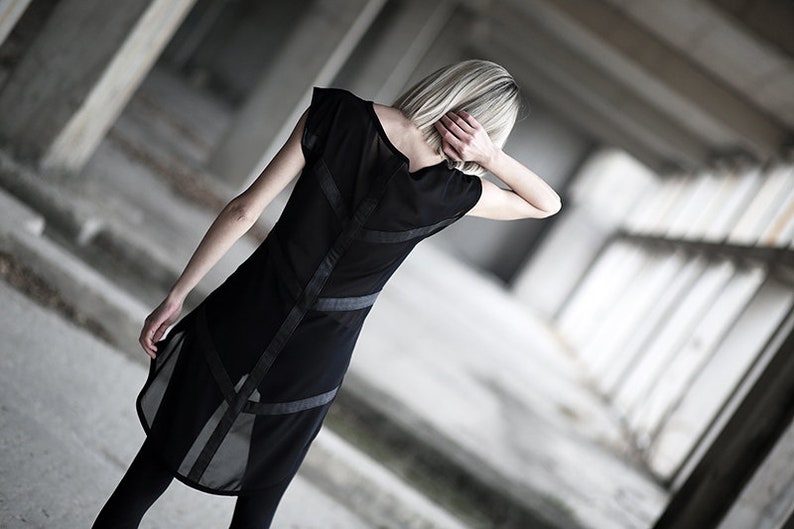 May include: Black dress with sheer panels and geometric accents. The dress features a sleeveless design with a high neckline and a flowing, asymmetrical hemline. The dress is paired with black leggings.