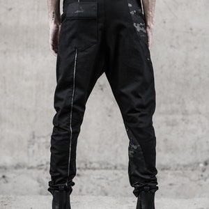 May include: Black pants with a unique design, featuring a drop-crotch style and a silver stripe down the side. The pants have a camouflage pattern on the side and back pocket. The model is wearing black boots. The pants are made of a durable material.