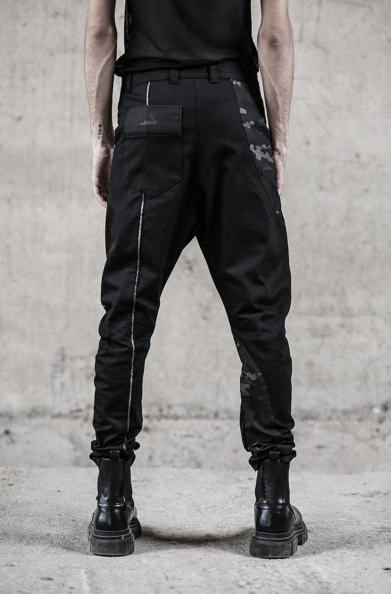 May include: Black pants with a unique design, featuring a drop-crotch style and a silver stripe down the side. The pants have a camouflage pattern on the side and back pocket. The model is wearing black boots. The pants are made of a durable material.