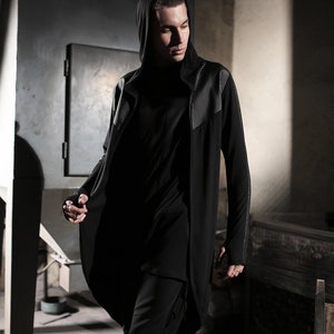 May include: A black hooded long coat with studded accents on the shoulders and sleeves. The coat is paired with a black top and cropped pants. The outfit is part of a modern, urban fashion collection.