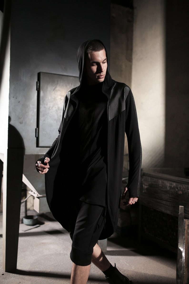 May include: A black hooded long coat with studded accents on the shoulders and sleeves. The coat is paired with a black top and cropped pants. The outfit is part of a modern, urban fashion collection.
