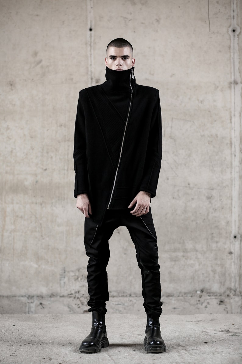 May include: A person wearing a black outfit with a high collar and a diagonal zipper. The outfit includes a long-sleeved top and matching pants. The person is also wearing black boots. The background is a concrete wall.
