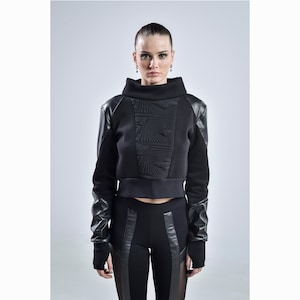 May include: A black crop top with a high collar and leather sleeves. The top features a textured panel on the front. The model is also wearing black leggings with leather panels. The outfit is a modern, edgy style.