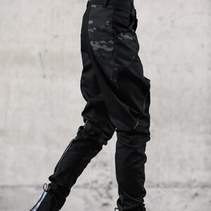 May include: Black pants with a camouflage pattern on the side, paired with black Chelsea boots. The pants have a slim fit and a subtle design. The boots have a chunky sole. The outfit is all black, creating a sleek, modern look.