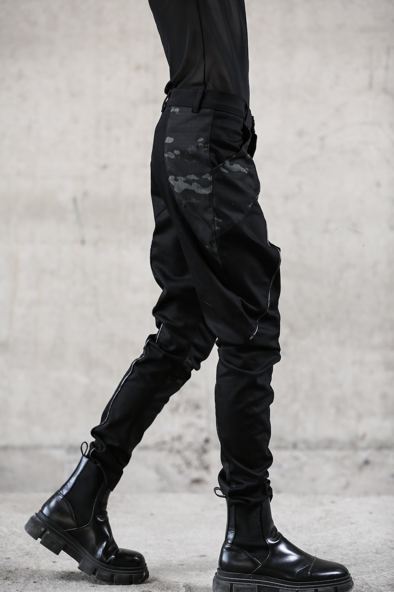 May include: Black pants with a camouflage pattern on the side, paired with black Chelsea boots. The pants have a slim fit and a subtle design. The boots have a chunky sole. The outfit is all black, creating a sleek, modern look.