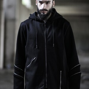 May include: A black zip-up hoodie with silver zipper accents and a drawstring hood. The hoodie has a long, asymmetrical hemline and a relaxed fit.