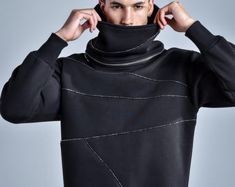 Puede incluir: Una sudadera negra de manga larga con cuello alto y una prominente cremallera plateada. La prenda incluye detalles de ribetes plateados en diagonal a través del pecho y el torso, con puños y dobladillo acanalados.