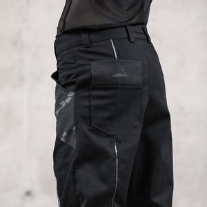 May include: Black pants with a unique design. The pants feature a large pocket on the back and a decorative design on the side. The pants have a silver trim along the seams. The pants are made of a dark material.