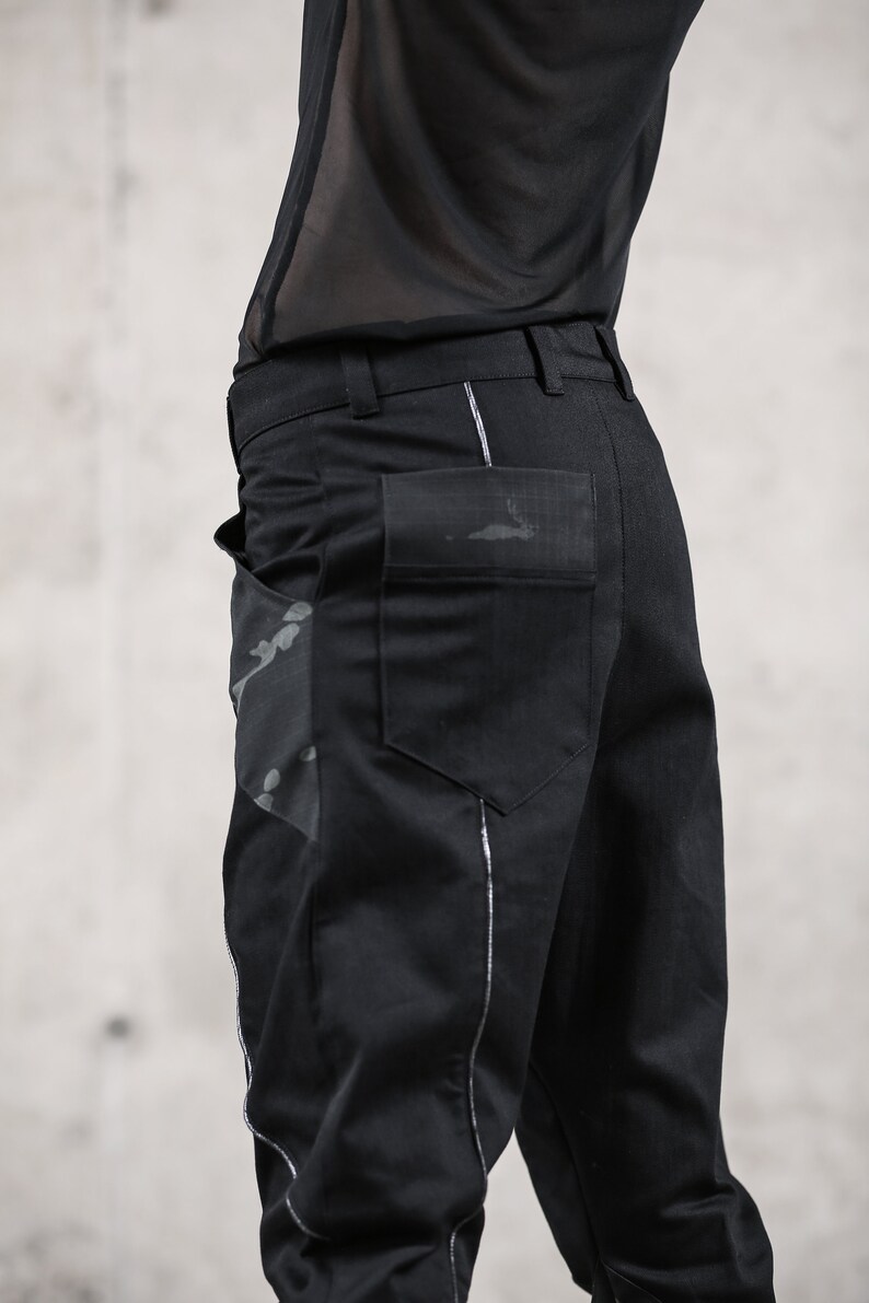 May include: Black pants with a unique design. The pants feature a large pocket on the back and a decorative design on the side. The pants have a silver trim along the seams. The pants are made of a dark material.