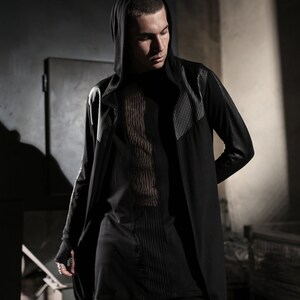 May include: Black hooded long coat with silver studded accents on the shoulders and sleeves. The coat is paired with a black top featuring vertical textured detailing. The overall style is dark and modern.