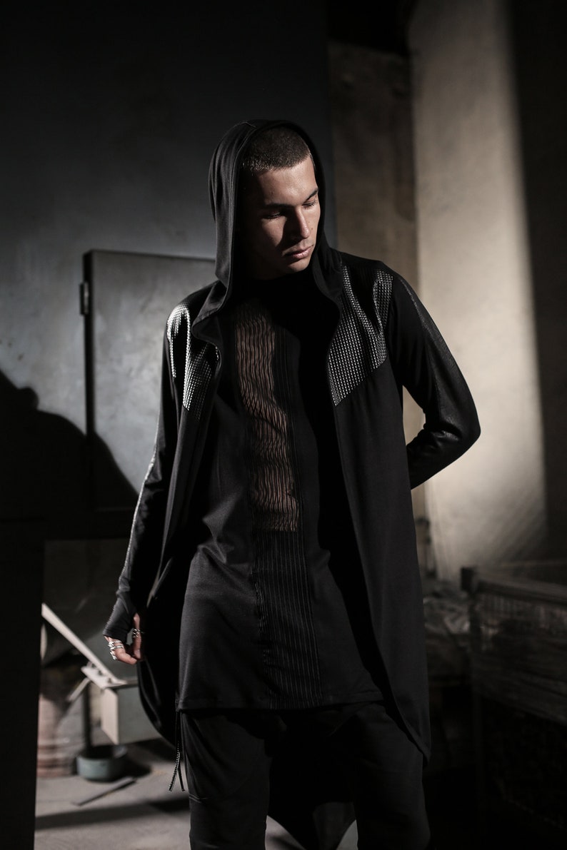 May include: Black hooded long coat with silver studded accents on the shoulders and sleeves. The coat is paired with a black top featuring vertical textured detailing. The overall style is dark and modern.