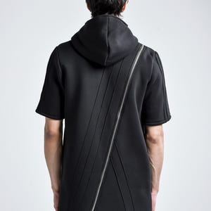 Puede incluir: Sudadera con capucha negra de manga corta con cremallera plateada diagonal. La sudadera tiene capucha y detalles de costura verticales en la parte delantera y trasera. El modelo est&aacute; de espaldas.