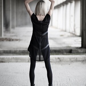 May include: A black dress with sheer panels and leather-like accents. The dress is paired with black leggings and ankle boots. The person's arms are raised above their head. The background is a concrete structure.
