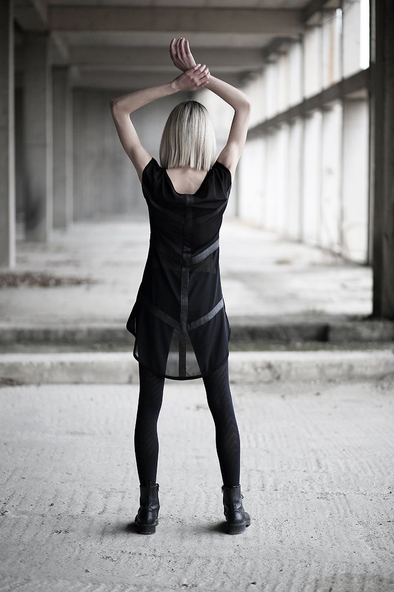 May include: A black dress with sheer panels and leather-like accents. The dress is paired with black leggings and ankle boots. The person's arms are raised above their head. The background is a concrete structure.