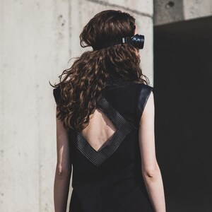 May include: A black sleeveless dress with a geometric design on the back. The dress has leather accents on the shoulders and back. The person is wearing a black headpiece and wrist cuffs. The background is a concrete wall.
