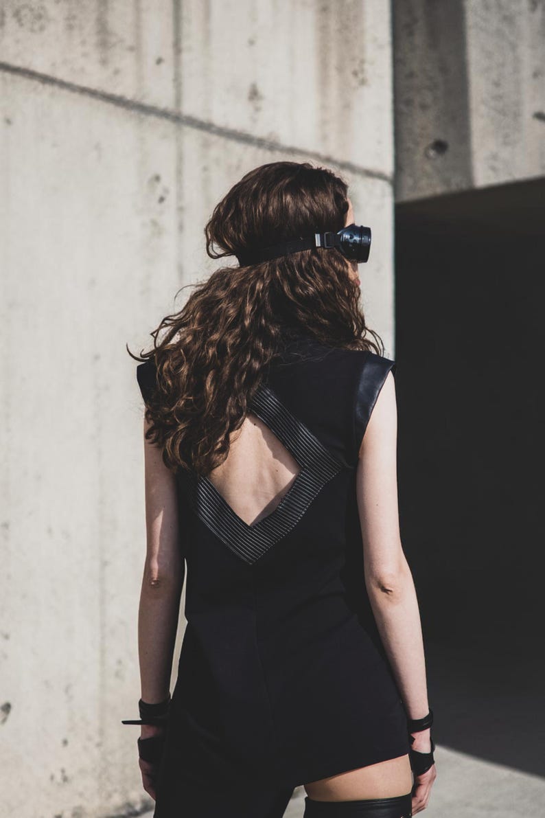 May include: A black sleeveless dress with a geometric design on the back. The dress has leather accents on the shoulders and back. The person is wearing a black headpiece and wrist cuffs. The background is a concrete wall.