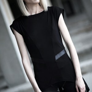 May include: A black top with a unique design, featuring a geometric panel and leather-like accents. The top has short sleeves and is paired with black leggings. The model has blonde hair and is in an outdoor setting.