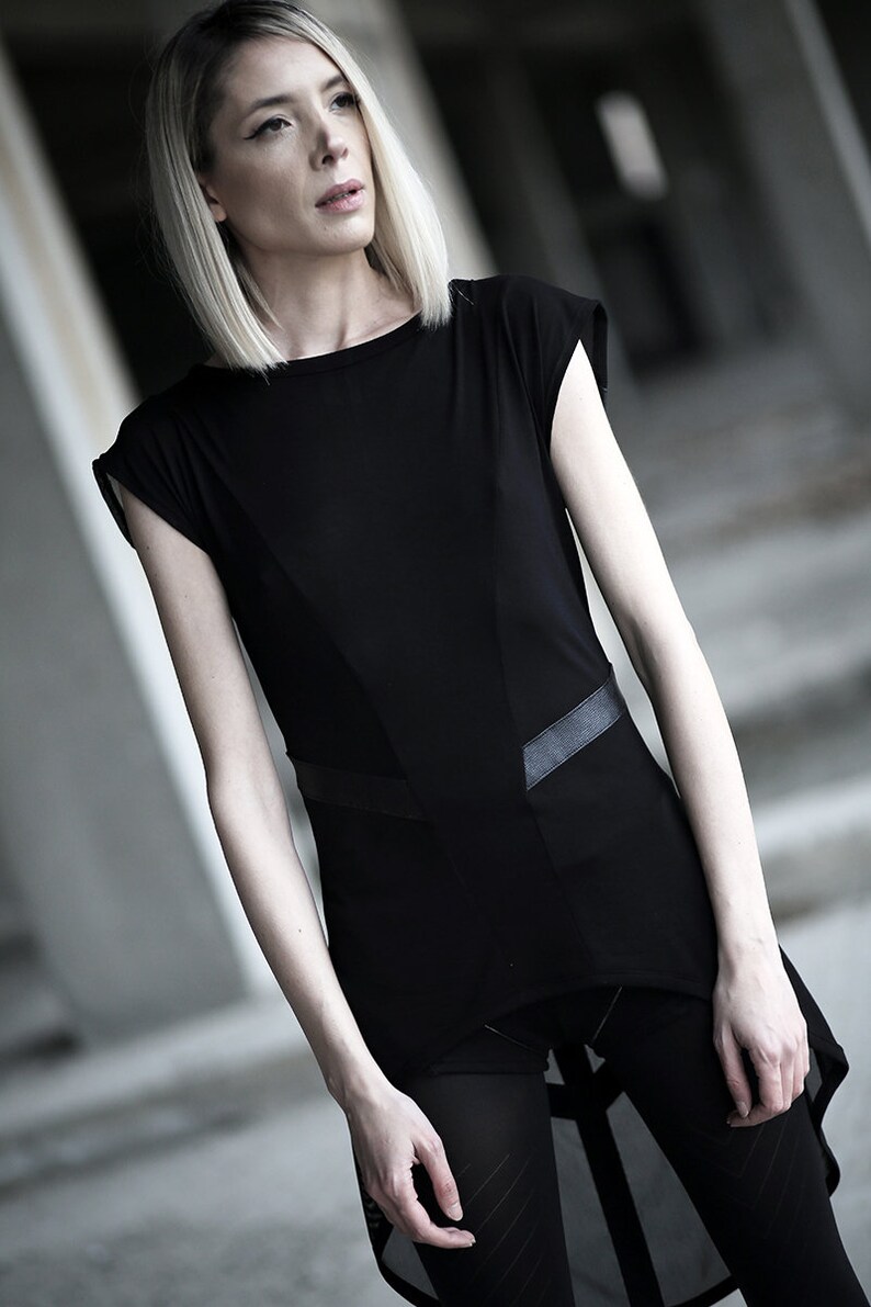 May include: A black top with a unique design, featuring a geometric panel and leather-like accents. The top has short sleeves and is paired with black leggings. The model has blonde hair and is in an outdoor setting.