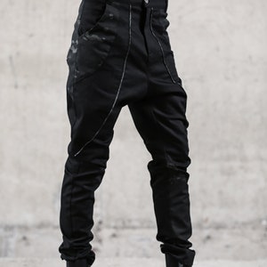 May include: Black pants with a unique design. The pants feature a high waist, a button closure, and pockets with a camouflage pattern. Silver stitching accents the seams. The pants are paired with black boots.