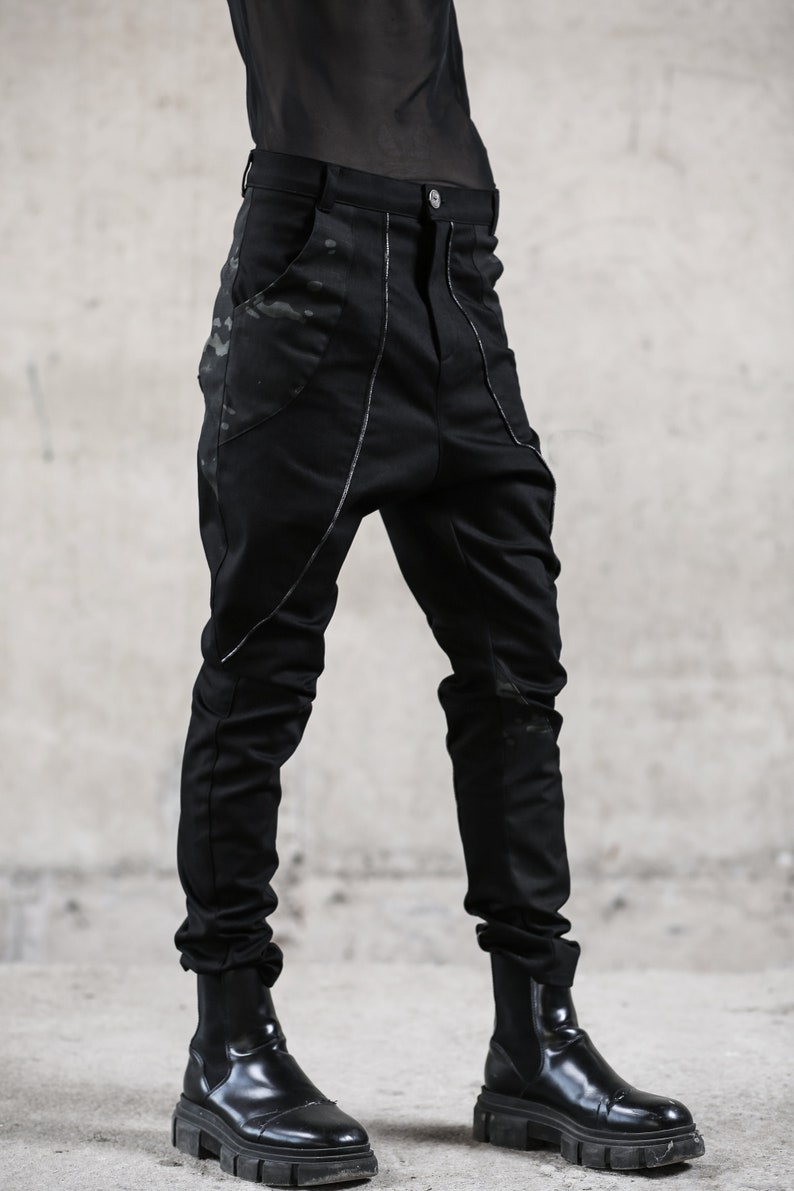 May include: Black pants with a unique design. The pants feature a high waist, a button closure, and pockets with a camouflage pattern. Silver stitching accents the seams. The pants are paired with black boots.