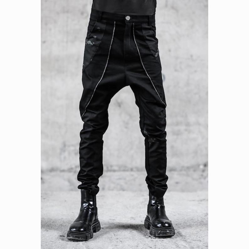 May include: Black pants with a unique design, featuring a low crotch and a tailored fit. The pants have white piping along the seams and are paired with black boots. The overall style is modern and edgy.