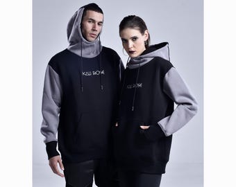 May include: Two people wearing black and gray hoodies. The hoodies have a high collar and a hood. The text "KALI ROSE" is embroidered on the chest. The hoodies have a kangaroo pocket.