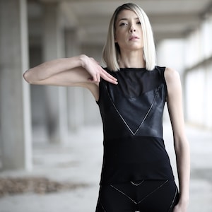 Op de afbeelding: Een vrouw met blond haar draagt een zwart, mouwloos topje met een lederen paneel en een transparante mesh-overlay. Het topje heeft een V-hals en een aansluitende pasvorm.