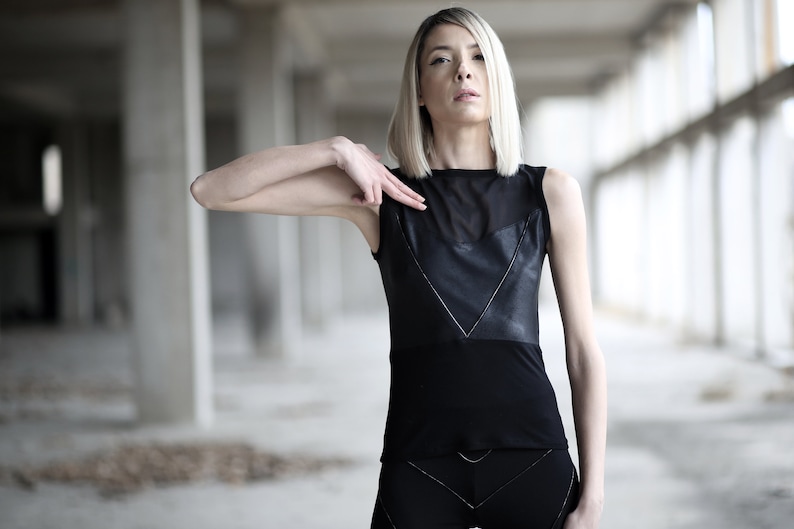 Op de afbeelding: Een vrouw met blond haar draagt een zwart, mouwloos topje met een lederen paneel en een transparante mesh-overlay. Het topje heeft een V-hals en een aansluitende pasvorm.