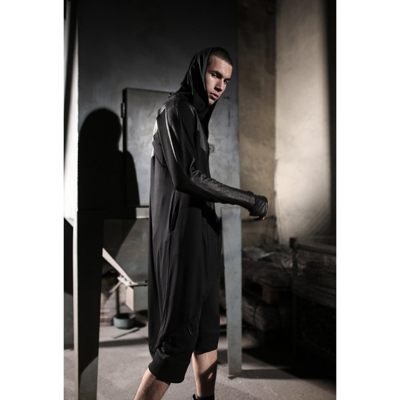 May include: A black hooded jumpsuit with a long, flowing design. The sleeves feature a textured pattern, and the garment has a relaxed fit. The model is wearing fingerless gloves. The background is a dimly lit industrial setting.