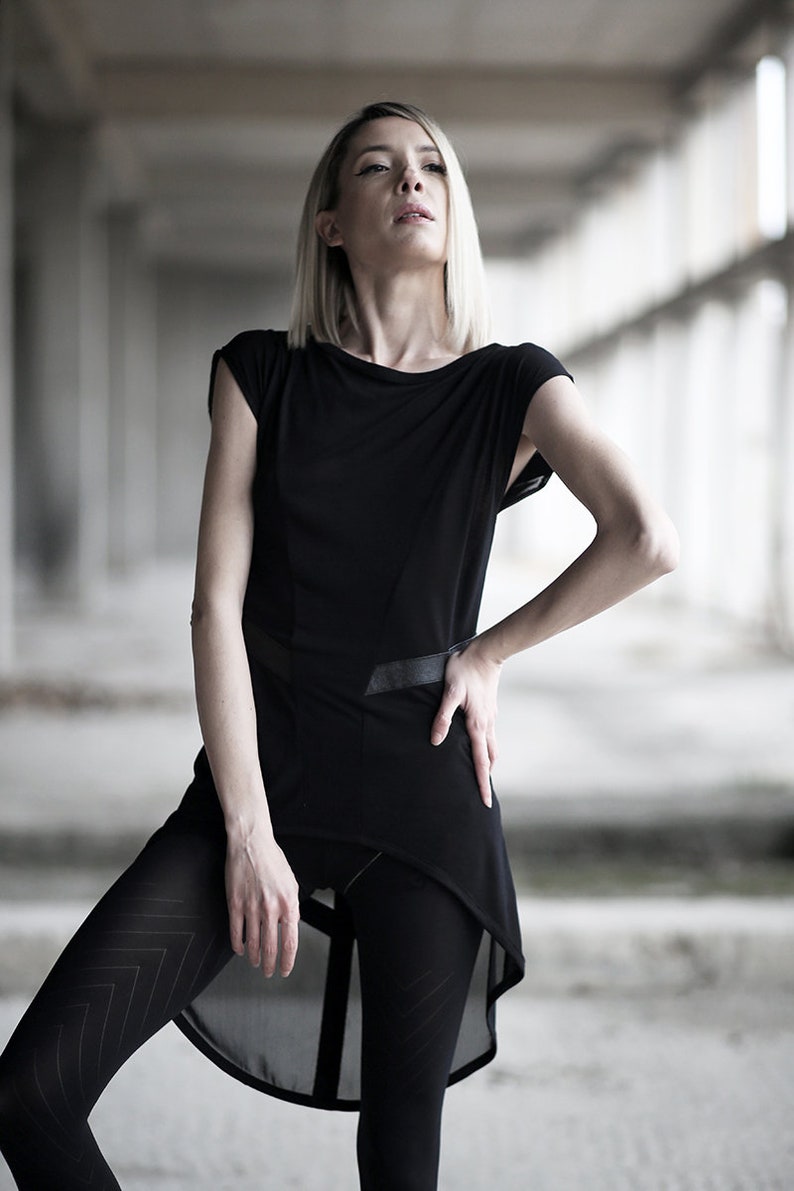 May include: A black asymmetrical tunic with a leather belt detail. The tunic has short sleeves and a flowing, layered design. The model is wearing black leggings with a chevron pattern. The background is a blurred, industrial setting.
