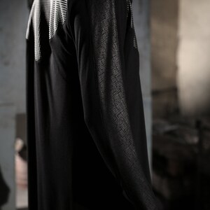 May include: Black hooded long-sleeve garment with textured accents on the shoulders and sleeves. The garment has a flowing, floor-length design. The shoulder area features a white, grid-like pattern. The sleeves have a textured, cross-hatch design.