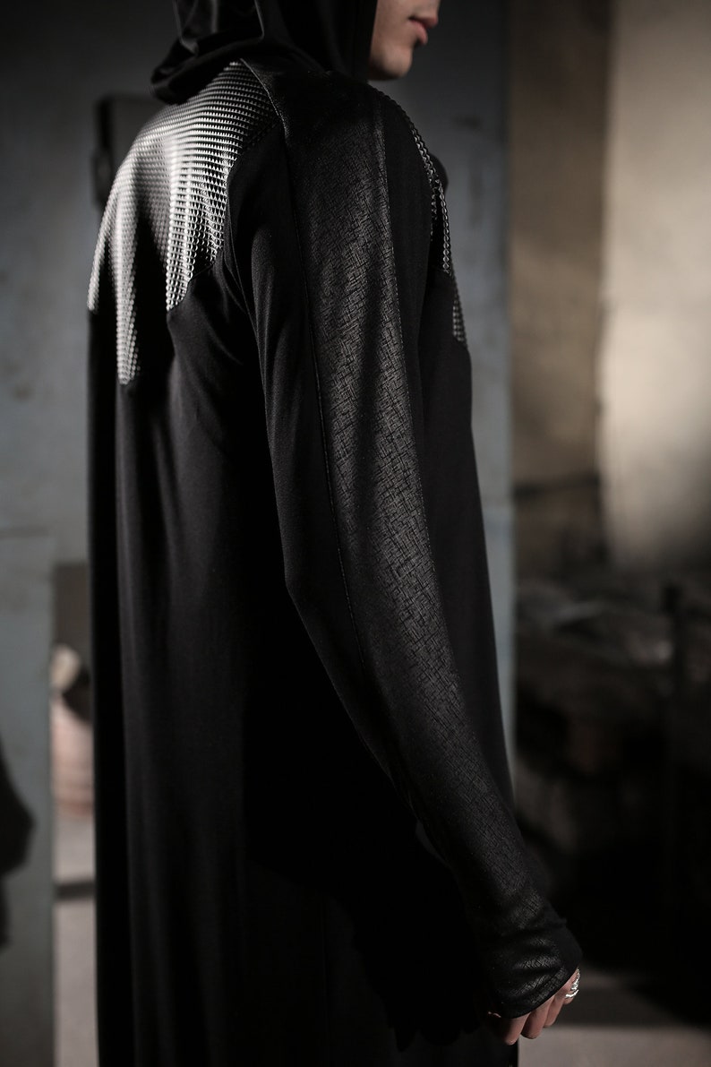 May include: Black hooded long-sleeve garment with textured accents on the shoulders and sleeves. The garment has a flowing, floor-length design. The shoulder area features a white, grid-like pattern. The sleeves have a textured, cross-hatch design.