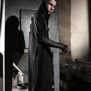 May include: A person wearing a black hooded jumpsuit with a unique design on the sleeves and back. The jumpsuit has a sleek, modern aesthetic, with a minimalist design. The person is standing in a dimly lit, industrial-style setting.