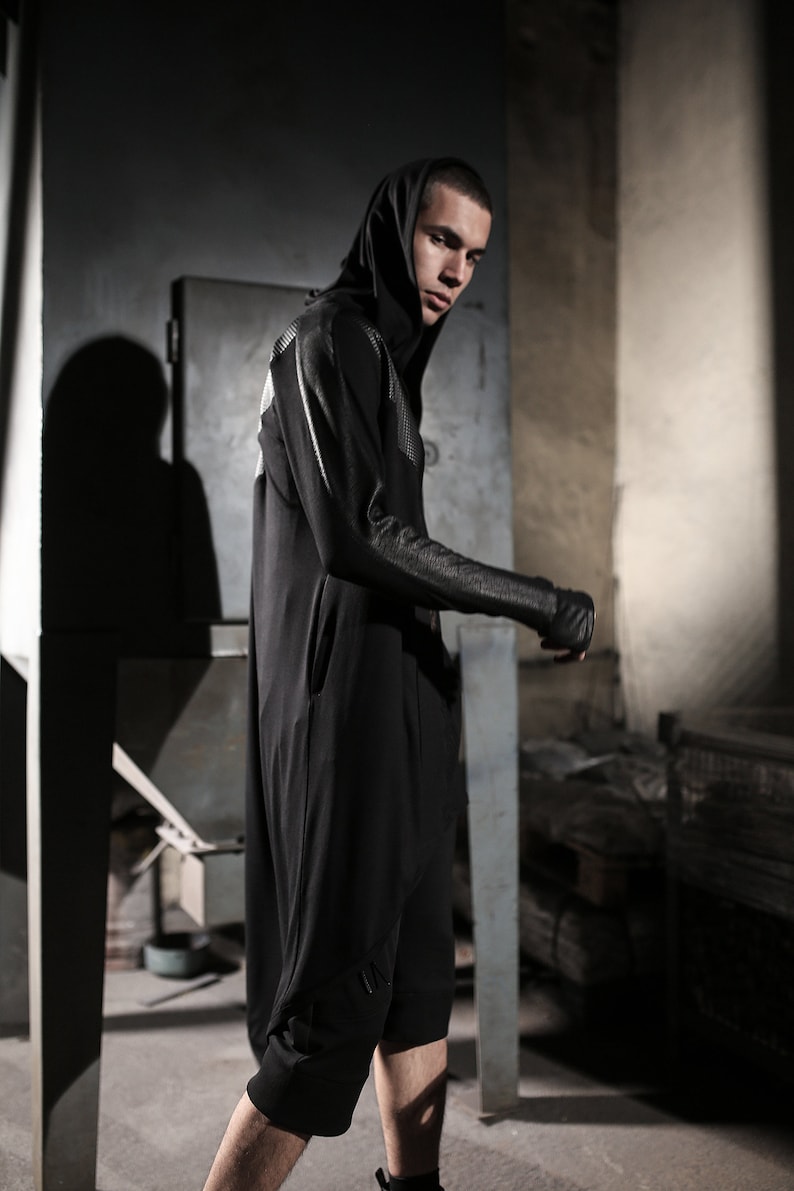 May include: A person wearing a black hooded jumpsuit with a unique design on the sleeves and back. The jumpsuit has a sleek, modern aesthetic, with a minimalist design. The person is standing in a dimly lit, industrial-style setting.