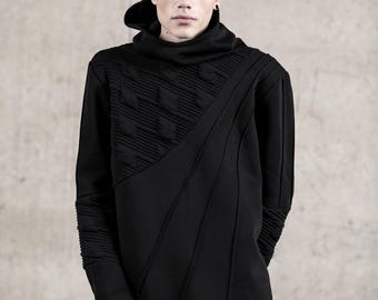 May include: A black long-sleeve hooded sweatshirt featuring a high collar, a textured diagonal panel with raised geometric patterns on the chest, and ribbed detailing on the sleeves and hem. The garment includes thumbhole cuffs.