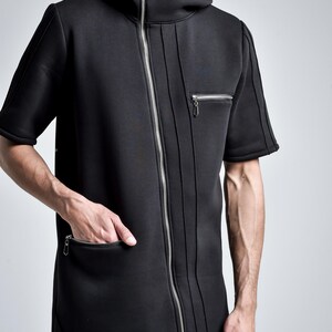 Puede incluir: Chaqueta negra con capucha y manga corta con cremallera plateada de largo completo. La chaqueta tiene un cuello alto, un peque&ntilde;o bolsillo con cremallera en el pecho y un bolsillo m&aacute;s grande en la parte delantera. Costuras verticales recorren la parte delantera de la chaqueta.