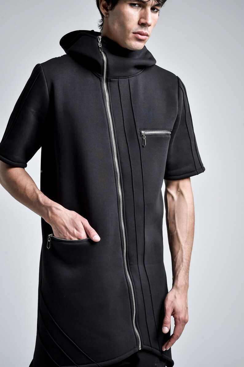 Puede incluir: Chaqueta negra con capucha y manga corta con cremallera plateada de largo completo. La chaqueta tiene un cuello alto, un peque&ntilde;o bolsillo con cremallera en el pecho y un bolsillo m&aacute;s grande en la parte delantera. Costuras verticales recorren la parte delantera de la chaqueta.