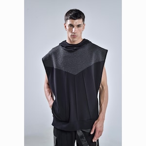 Puede incluir: Sudadera sin mangas negra con un patrón geométrico gris texturizado en los hombros y el pecho. La sudadera con capucha tiene una capucha y un bolsillo de canguro. El modelo lleva pantalones negros.
