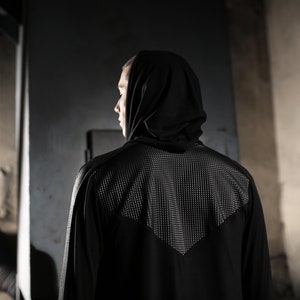 May include: A black hooded jacket with a textured panel on the upper back. The jacket has long sleeves and a sleek, modern design. The person is facing away from the camera, with the hood up.