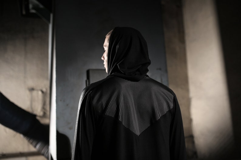 May include: A black hooded jacket with a textured panel on the upper back. The jacket has long sleeves and a sleek, modern design. The person is facing away from the camera, with the hood up.