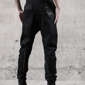 May include: Black pants with a unique design, featuring a drop-crotch style and a subtle camouflage pattern on the pockets and side panels. The pants have a silver stripe down the side and are paired with black boots.