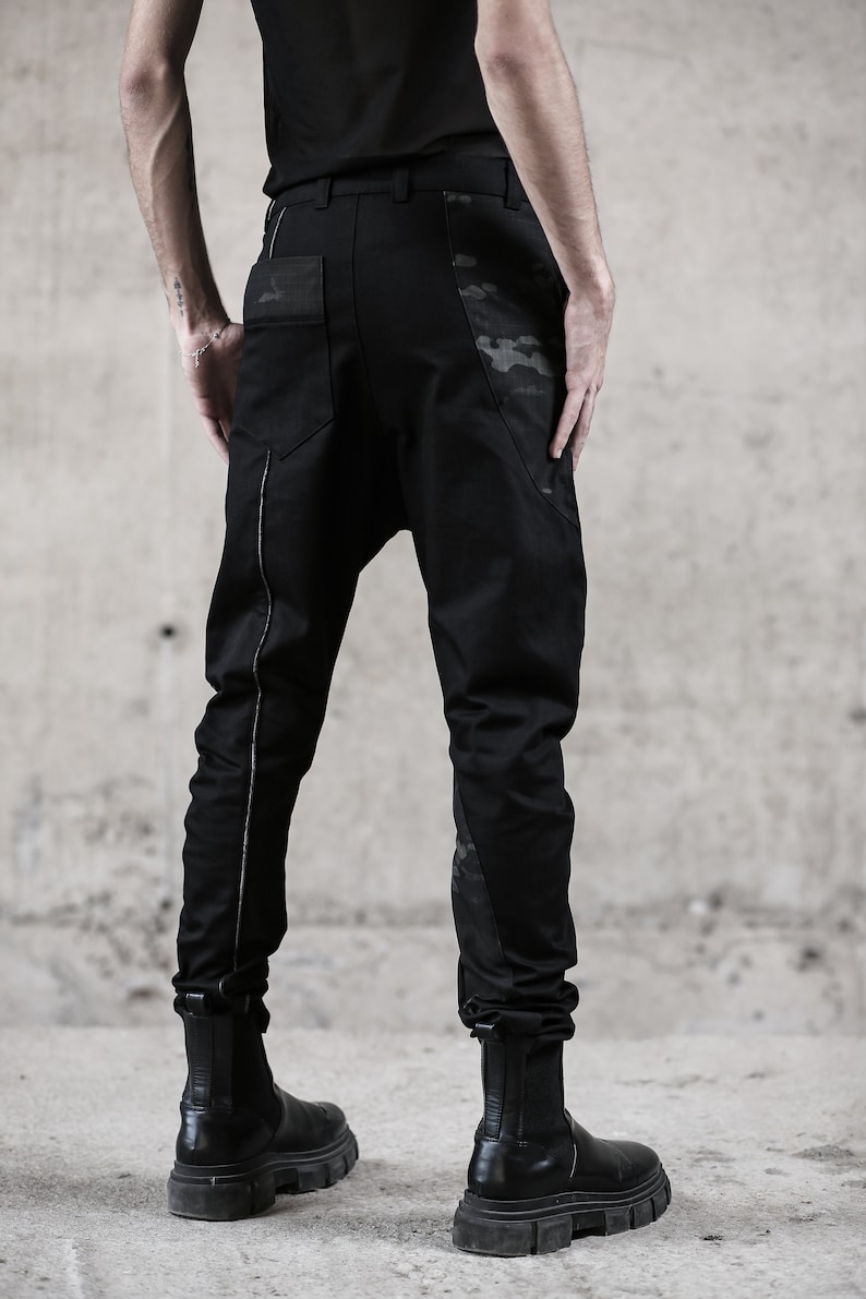 May include: Black pants with a unique design, featuring a drop-crotch style and a subtle camouflage pattern on the pockets and side panels. The pants have a silver stripe down the side and are paired with black boots.