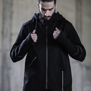 May include: A black zip-up hoodie with a long, pointed hemline and a hood. The hoodie has a zipper closure and two zippered pockets on the front. The sleeves have a unique design with a zippered cuff that can be worn as a thumbhole.
