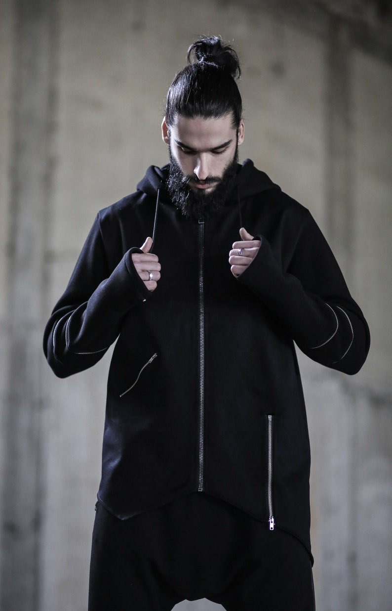 May include: A black zip-up hoodie with a long, pointed hemline and a hood. The hoodie has a zipper closure and two zippered pockets on the front. The sleeves have a unique design with a zippered cuff that can be worn as a thumbhole.
