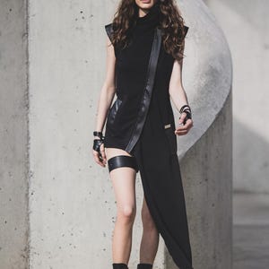 May include: A black asymmetrical dress with leather accents and a high collar. The dress features a long, flowing panel and is paired with black boots and a leg garter. The model is wearing dark sunglasses and fingerless gloves.