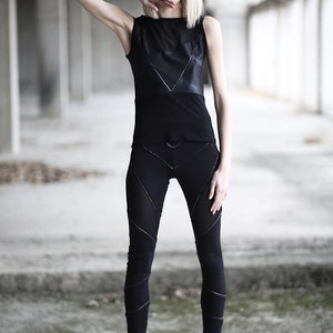 Op de afbeelding: Een vrouw draagt een zwart tanktop met een lederen paneel en zwarte leggings met geometrische stiksels. Ze draagt zwarte combatlaarzen en staat in een betonnen gebouw.